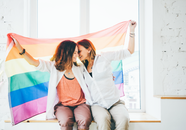 two women sitting on a window sill holding a rainbow flag