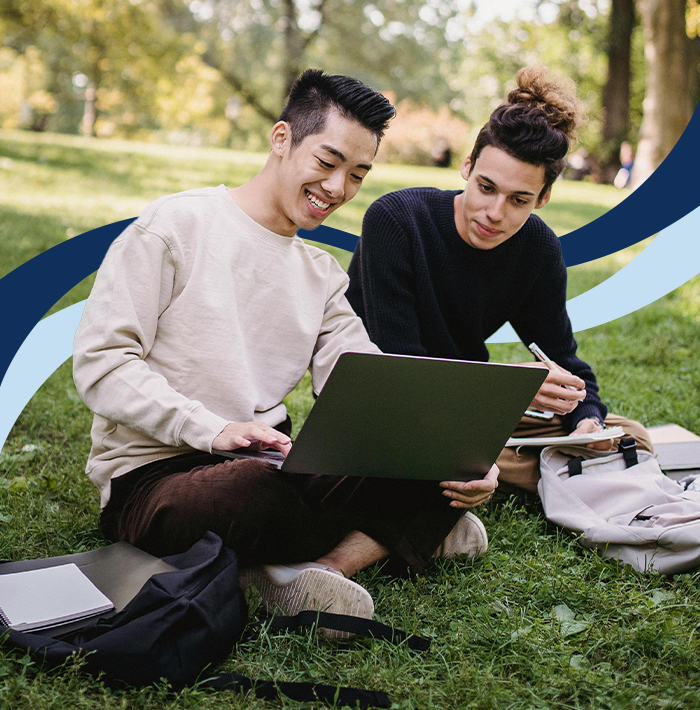 two men sitting on the grass looking at a laptop